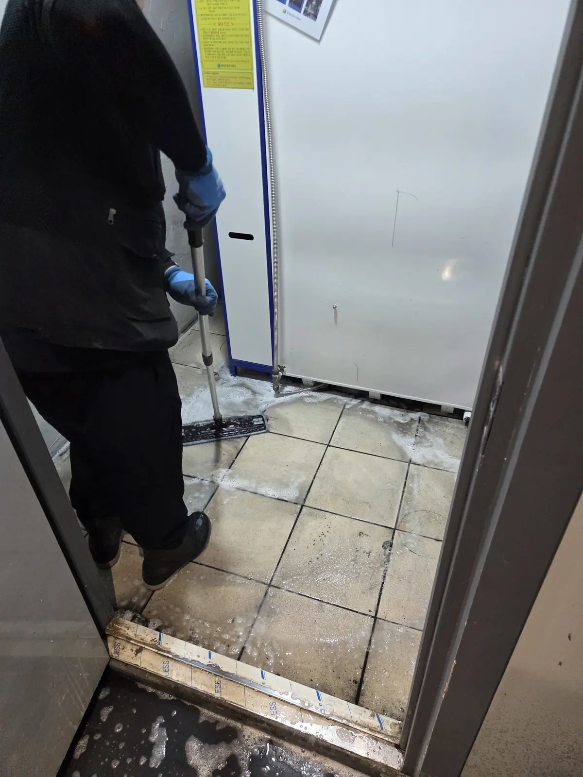 지하 출입구 타일 바닥에 세제를 풀고 밀대로 문질러 세정하는 작업자 — 줄눈에 남은 오염을 걷어내는 단계