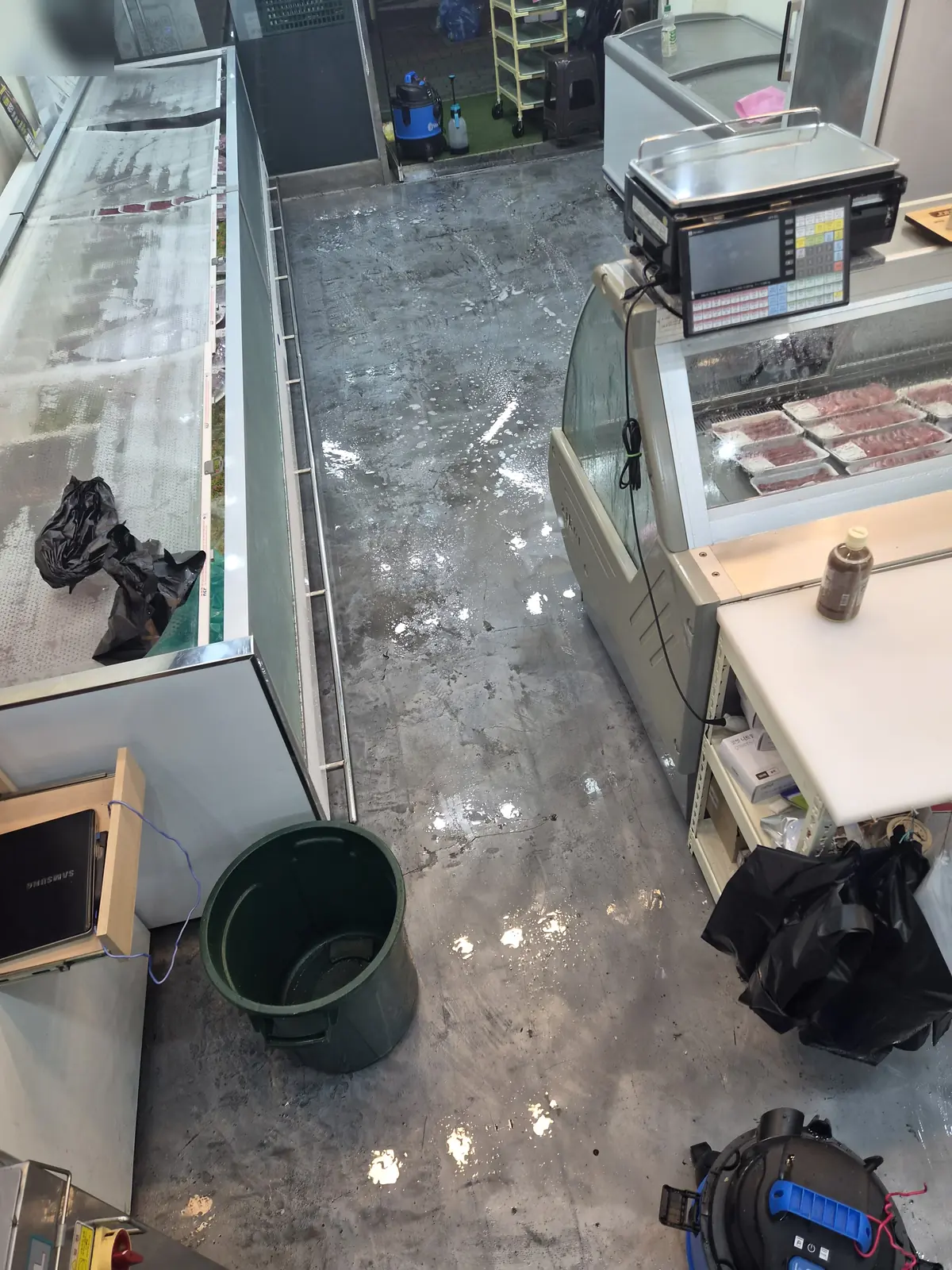 위에서 내려다본 정육점 통로 — 진열장 사이 바닥 전체가 젖어 물기가 번진 범위가 드러난 상태