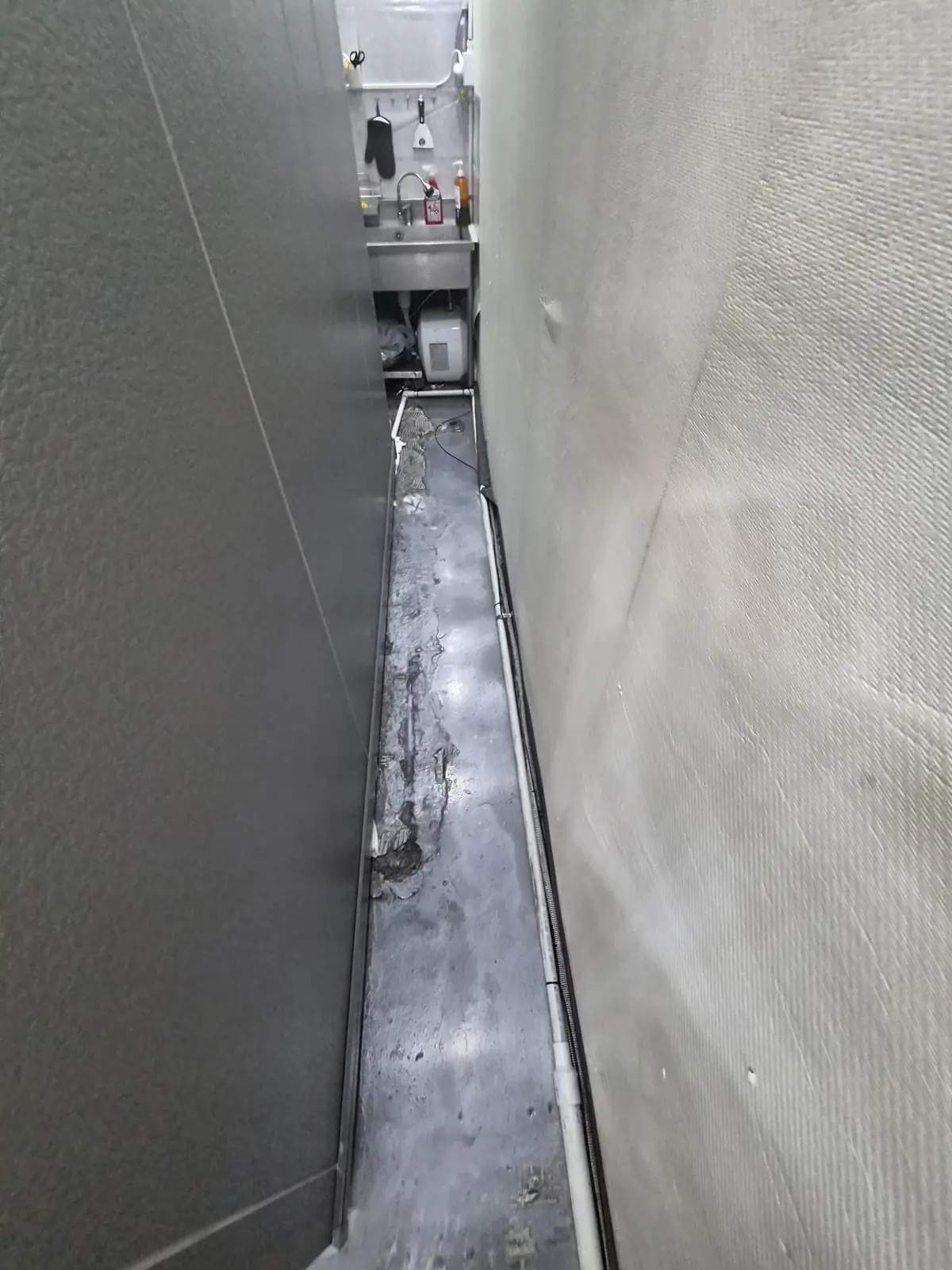 벽 사이 좁은 통로를 따라 배관이 지나가고 바닥 마감이 파손된 구간 — 오수가 지나간 자리