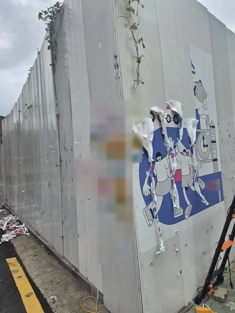 공사장 가림막 모서리에 붙은 그림 시트가 여러 줄로 말려 벗겨지는 모습과 옆에 세워 둔 사다리