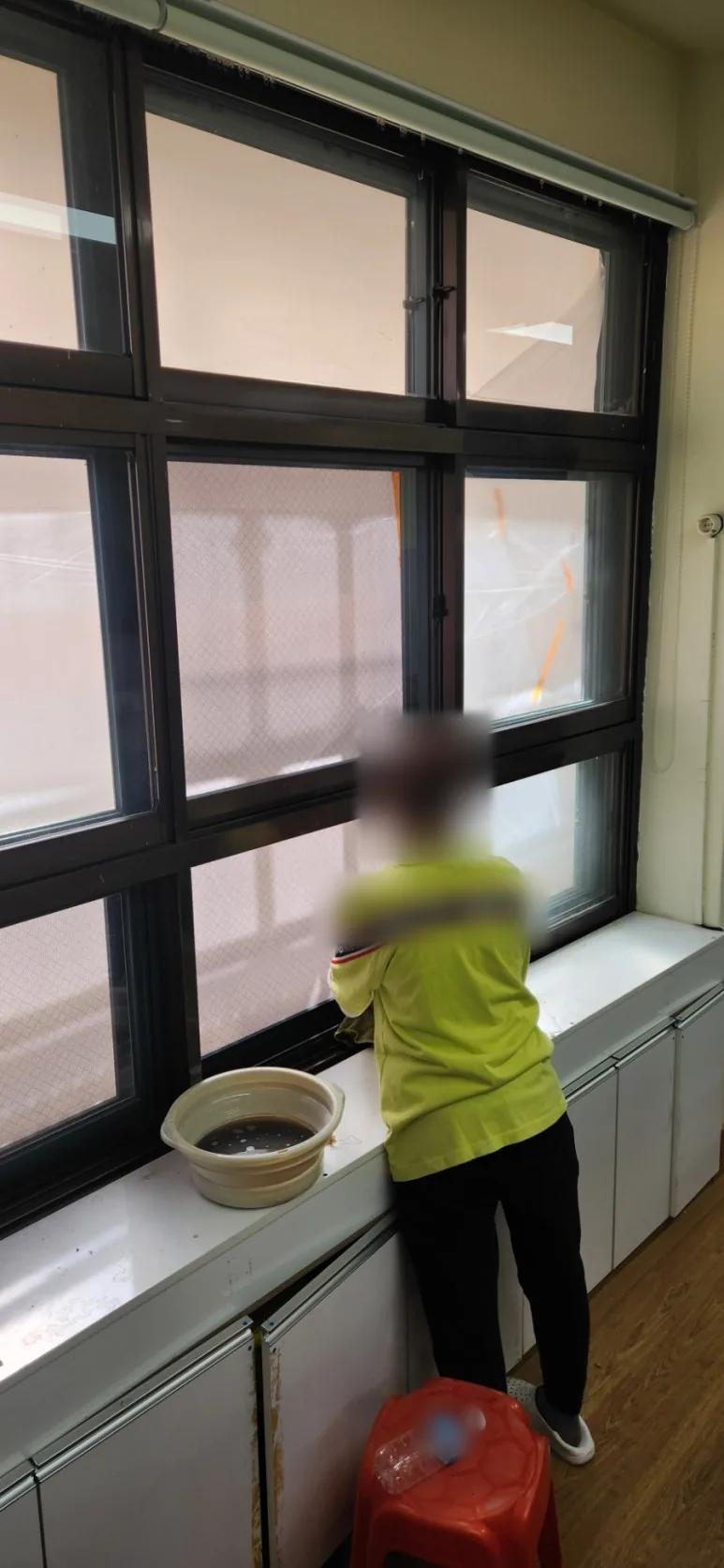 교실 창가 수납장 위에 물 대야를 올려 두고 작업자가 여러 칸으로 나뉜 큰 유리창을 안쪽에서 닦는 모습