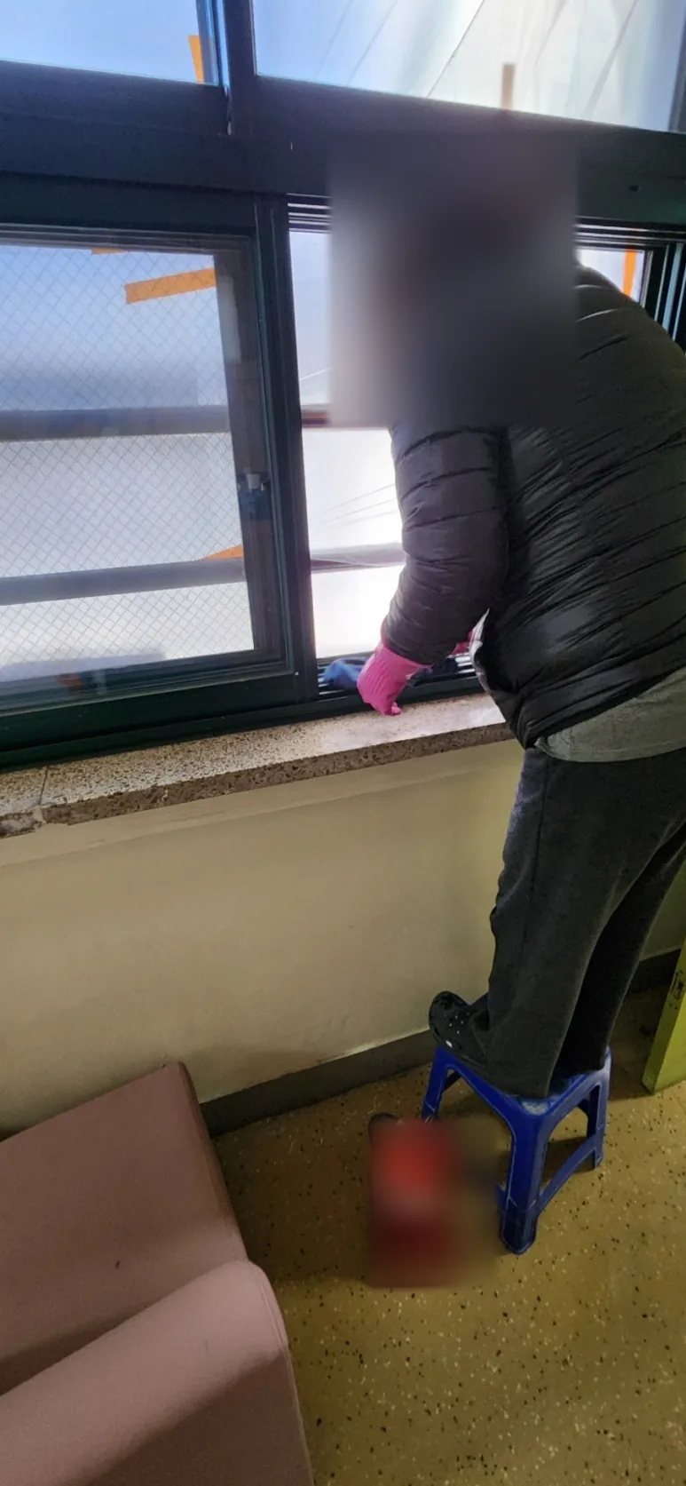 파란 발판에 올라선 작업자가 분홍 장갑을 끼고 학교 복도 창문 아래 레일과 창틀을 걸레로 닦는 모습